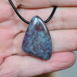 May include: A triangular pendant with a blue and maroon stone, hanging from a black cord. The stone displays a mottled pattern with shades of blue and maroon. The pendant has a smooth, polished finish.