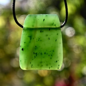 Könnte beinhalten: Ein grüner Jade-Anhänger mit dunkelgrünen Flecken, der an einer schwarzen Schnur hängt.