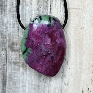 Op de afbeelding: Een roze en groene edelsteen hanger aan een zwart koord. De hanger heeft een natuurlijke, onregelmatige vorm met een glad, gepolijst oppervlak.