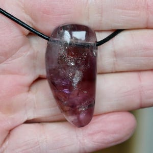 Könnte beinhalten: Ein durchscheinender, tropfenförmiger Anhänger in dunklem Lila, an einer schwarzen Kordel befestigt. Der Stein weist interne Einschlüsse und eine polierte Oberfläche auf und bildet eine einzigartige, natürliche Edelsteinkette.