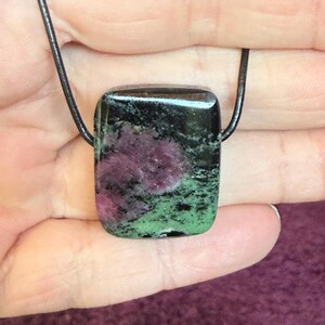 Op de afbeelding: Een vierkante hangerketting met een gepolijste steen. De steen heeft een zwarte bovenkant, een centraal gedeelte van diep roze en groen aan de onderkant. Hij is geregen aan een zwart koord.