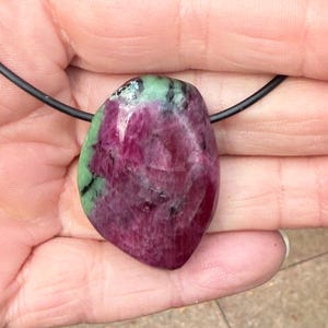 Peut inclure: Un pendentif en pierre précieuse poli avec un cordon noir. Le pendentif présente un mélange de couleurs, avec une base rouge-violet foncé et des taches vertes. La pierre a une forme lisse et arrondie, et est tenue par un cordon noir.