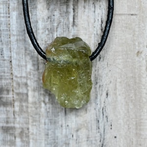 Könnte beinhalten: Ein grüner Kristall-Anhänger an einem schwarzen Lederband. Der Kristall ist roh und hat eine natürliche, unregelmäßige Form.
