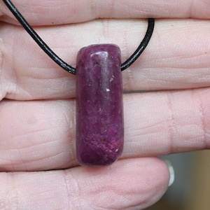 May include: A polished, rectangular pendant made of a deep magenta-coloured stone. The stone is suspended from a black cord necklace. The pendant is approximately 4 cm long and has a smooth, rounded top.