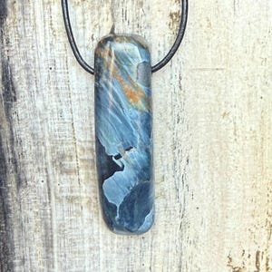 Könnte beinhalten: Ein blauer und brauner Edelsteinanhänger an einer schwarzen Kordel. Der polierte Stein hat eine rechteckige Form mit abgerundeten Kanten. Die Farben des Steins umfassen Blautöne, Braun und Weiß. Der Anhänger hängt vor einem hellen Holzgrund.