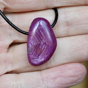 May include: A purplish-red gemstone pendant with a geometric pattern, suspended from a black cord. The stone's surface reflects light, showcasing its colour variations. The pendant is a unique piece of jewellery.
