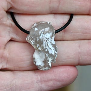 Könnte beinhalten: Ein klarer, unregelmäßig geformter Anhänger mit weißen Einschlüssen, der an einer schwarzen Kordel hängt. Der Anhänger wird in einer Hand gehalten und zeigt seine einzigartige, organische Form. Die Halskette ist ein Schmuckstück.