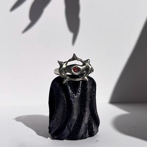 以下が含まれることがあります： 鋭い尖った要素と小さな赤い丸い石が特徴の、ユニークなデザインのシルバーリング。 リングは、白い背景に対して、質感のある黒いスタンドに展示されており、上には爪のような影があります。