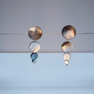 Peut inclure: Une paire de boucles d'oreilles pendantes avec un design superposé. Les boucles d'oreilles présentent quatre disques ronds, irisés, en nacre dans des tons de gris, blanc et bleu. Les disques sont reliés par du métal doré.