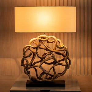 May include: A table lamp featuring a rectangular beige lampshade. The base is a sphere constructed from interwoven, light brown branches. The lamp rests on a black base and a grey surface, set against a wooden panel background.