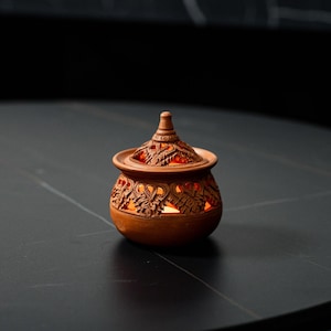 Op de afbeelding: Een terracotta-kleurige, ingewikkeld gesneden keramische pot met een deksel. Het ontwerp van de pot heeft bloempatronen en wordt van binnenuit verlicht, waardoor een warme gloed ontstaat. De pot is een decoratief item.
