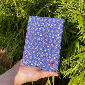 Pode incluir: Um caderno retangular revestido de tecido com fundo azul e um padrão repetitivo de pequenas flores laranja e azul-claras. Um único botão pequeno de cor vermelha brilhante em forma de coração está fixado no canto inferior direito.