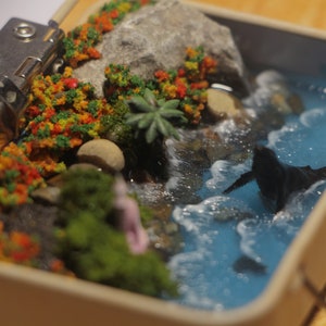 Puede incluir: Una escena de diorama en miniatura con una pequeña cascada que desemboca en un océano azul con espuma blanca. La escena está ambientada en una pequeña caja de hojalata con tapa con bisagras. Hay rocas verdes y marrones, musgo verde y follaje rojo y verde.