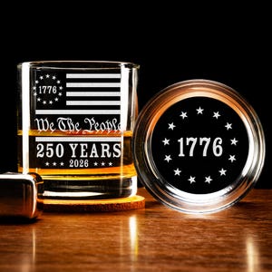 Puede incluir: Un vaso de whisky transparente con el dise&ntilde;o de la bandera estadounidense, el texto "1776", "We The People", "250 YEARS" y "2026". Tambi&eacute;n se ven un posavasos y una tapa a juego con el texto "1776" y estrellas. El vaso est&aacute; lleno de l&iacute;quido &aacute;mbar.