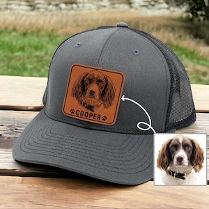 May include: A gray and black trucker hat with a brown leather patch featuring a dog's portrait and the name "COOPER".