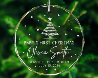 May include: A clear glass Christmas ornament with a white snowflake design and the text "BABY'S FIRST CHRISTMAS Olivia Smith 7LBS 8OZ | 19 IN | 11:30 AM JULY 15, 2024".