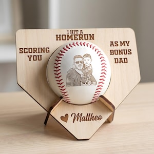May include: A light-colored wooden home plate display stand holding a white baseball with red stitching. The baseball features a laser-engraved portrait of two people. The stand is engraved with the text I HIT A HOMERUN SCORING YOU AS MY BONUS DAD and a heart icon with the name Mattheo.
