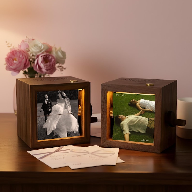 May include: Two wooden photo cubes with illuminated images of a wedding and a couple on grass. The cubes have a dark wood finish, gold accents, and a small crank. A bouquet of pink and white flowers sits behind the cubes.