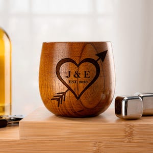 May include: A wooden stemless wine glass with a heart and arrow design. The heart contains the initials "J & E" and the year "EST. 2020". The glass is sitting on a wooden surface, with stainless steel ice cubes in the background.