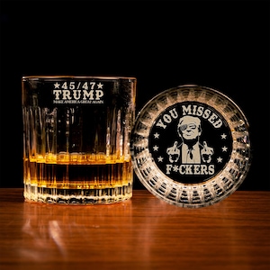 May include: A whiskey glass with the text "45/47 TRUMP MAKE AMERICA GREAT AGAIN" and a coaster with the text "YOU MISSED F*CKERS" and an image of a person. The glass is filled with amber liquid.