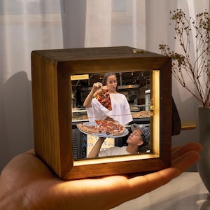 May include: A wooden music box with a photo of a pizza restaurant scene. The photo shows a woman holding a slice of pizza and a man holding a pizza. The text "TAKEAWAY" is visible. The box is held in a hand.