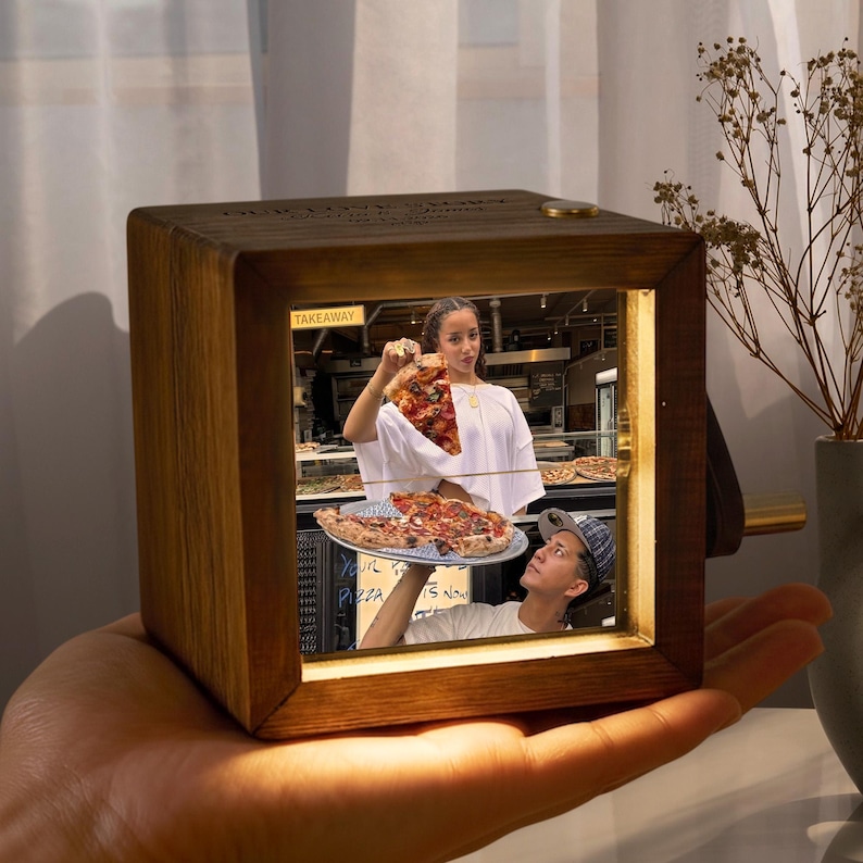 May include: A wooden music box with a photo of a pizza restaurant scene. The photo shows a woman holding a slice of pizza and a man holding a pizza. The text "TAKEAWAY" is visible. The box is held in a hand.