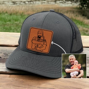 May include: A black and grey trucker hat with a brown leather patch featuring a line drawing of a person holding two children. The patch is on the front of the hat.