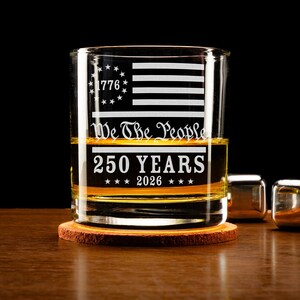 Puede incluir: Un vaso transparente con un dise&ntilde;o patri&oacute;tico. El vaso presenta una bandera estadounidense grabada, el texto "1776", "We The People" y "250 YEARS 2026". El vaso est&aacute; sobre un posavasos de madera, con cubitos de hielo met&aacute;licos cerca.