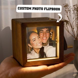 May include: A wooden custom photo flipbook with a photo of a smiling couple. The flipbook is square-shaped with a light brown finish and a small metal handle. The text "CUSTOM PHOTO FLIPBOOK" is in a white speech bubble.