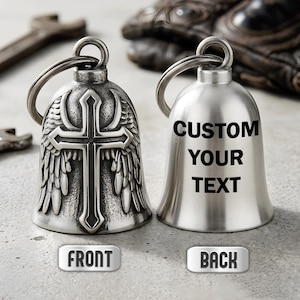 May include: Two silver-toned metal bells with split rings. The left bell features an embossed cross with wings in a dark, weathered finish. The right bell has a smooth, brushed finish with the black text CUSTOM YOUR TEXT printed on it.