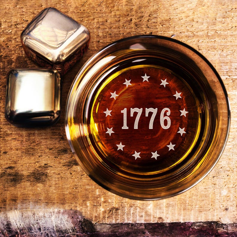 Puede incluir: Un vaso con l&iacute;quido &aacute;mbar con el a&ntilde;o "1776" y estrellas grabadas en el fondo. Dos cubitos de hielo met&aacute;licos est&aacute;n colocados junto al vaso sobre una superficie de madera. El vaso tiene una forma redonda cl&aacute;sica.