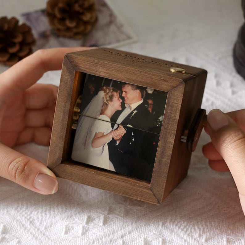 May include: A wooden photo cube with a framed picture of a couple dancing. The cube has a crank on the side and a small gold-colored accent on top. The wood has a warm, brown tone.