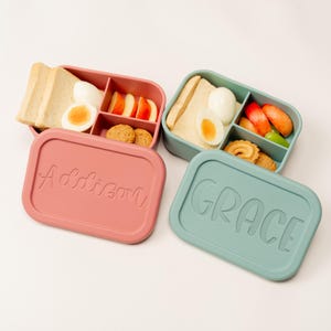 May include: Two personalized lunch boxes, one pink and one teal, each filled with food. The pink box lid says "Addison", and the teal box lid says "GRACE". The boxes contain bread, eggs, fruit, and cookies.