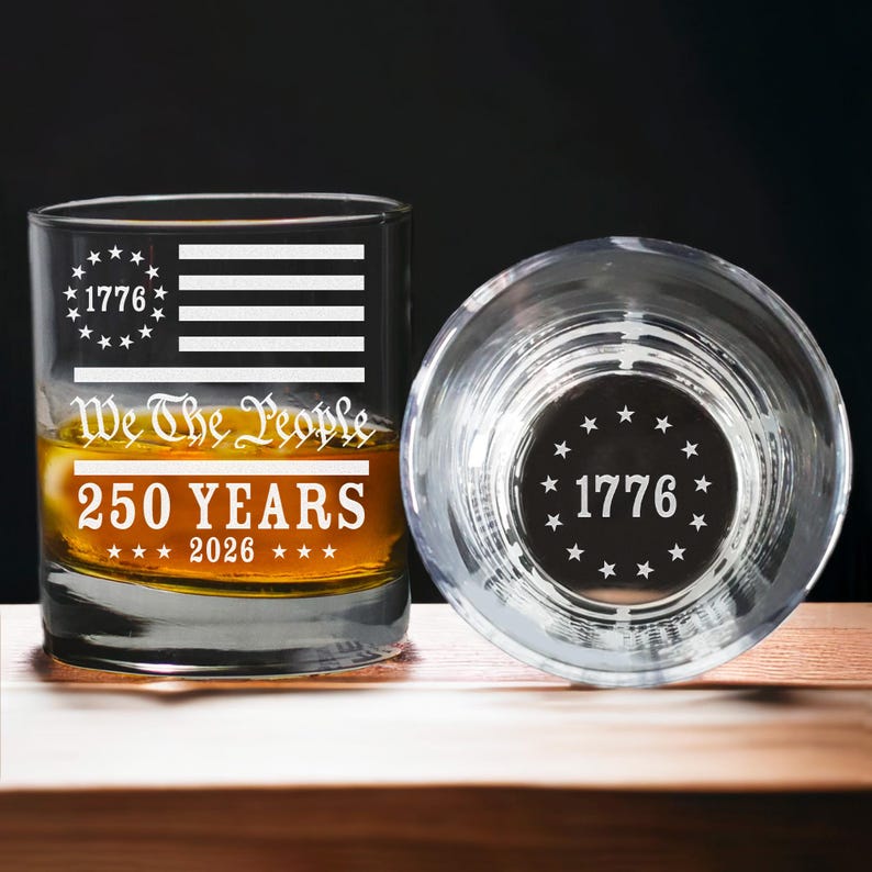 Puede incluir: Dos vasos de whisky transparentes con dise&ntilde;os patri&oacute;ticos. Uno presenta un dise&ntilde;o de bandera estadounidense blanca con el texto "1776", "We The People" y "250 YEARS 2026". El otro vaso tiene el texto "1776" y estrellas.