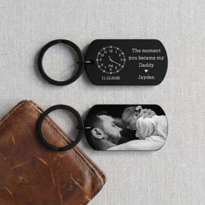 May include: Two black keychains with silver key rings. One keychain has a clock design and the text "The moment you became my Daddy" and the name "Jayden". The other keychain has a black and white photo of a father and baby.
