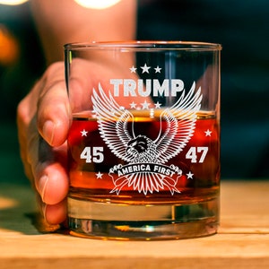 May include: A clear, lowball whiskey glass filled with amber liquid. The glass features the word "TRUMP" above an eagle design with the text "AMERICA FIRST" and the numbers "45" and "47". The glass is held by a person.