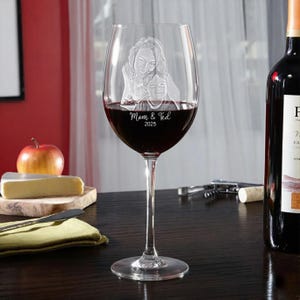 Pode incluir: Uma ta&ccedil;a de vinho transparente cheia de vinho tinto, com uma imagem gravada de uma m&atilde;e e uma crian&ccedil;a. As palavras "Mom & Ted 2025" est&atilde;o gravadas no vidro. Uma garrafa de vinho, uma ma&ccedil;&atilde; e queijo est&atilde;o no fundo.