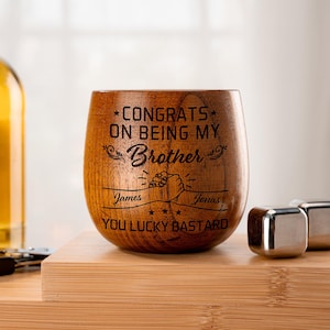 May include: A wooden stemless wine glass with the text "CONGRATS ON BEING MY Brother" and "YOU LUCKY BASTARD" engraved on it. The names "James" and "Jonas" are also engraved. Stainless steel ice cubes are next to the glass.