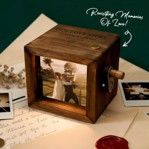 May include: A wooden photo cube with a picture of a couple. The cube has the text "OUR LOVE STORY" and the date "09.14.2026" engraved on it. The cube also has a crank and a small gold-colored button. The text "Revisiting Memories Of Love!" is written in cursive.