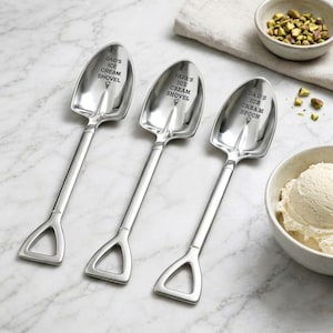 May include: Three silver ice cream shovels with the text "DAD'S ICE CREAM SHOVEL", "PAPA'S ICE CREAM SHOVEL", and "DAD'S ICE CREAM SPOON". Each shovel has a shovel-shaped scoop and a handle with a triangular opening. A bowl of ice cream and pistachios are in the background.