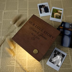 May include: A wooden box with the words "OUR LOVE STORY" engraved on the lid, along with the names "Keira & James" and the date "09.14.2026". The box is brown and has a gold-colored button. Three Polaroid photos are scattered around the box.