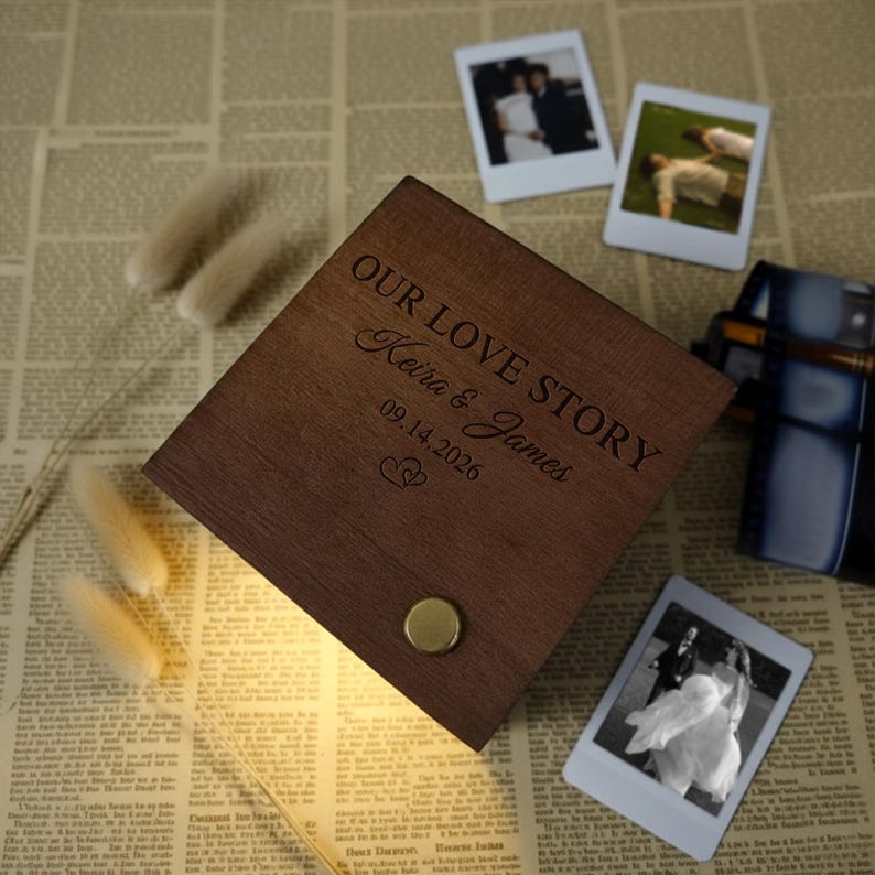 May include: A wooden box with the words "OUR LOVE STORY" engraved on the lid, along with the names "Keira & James" and the date "09.14.2026". The box is brown and has a gold-colored button. Three Polaroid photos are scattered around the box.