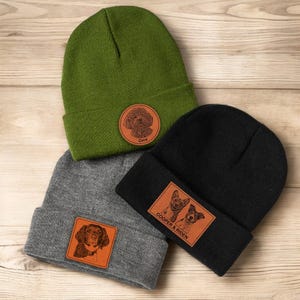 May include: Three knit beanies with personalized leather patches featuring dog portraits. The beanies are in green, black, and gray. The patches feature the names "Coco", "Cooper & Riden".