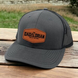 May include: A gray and black trucker hat with a brown leather patch that says "The Dadalorian This is the Way".
