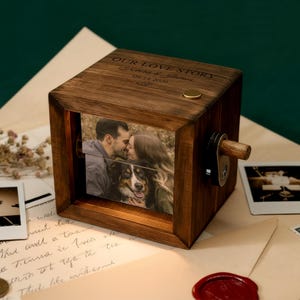 May include: A wooden photo cube with a rotating mechanism, displaying a couple kissing and a dog. The top of the cube is engraved with "OUR LOVE STORY" and a date. The cube is made of dark wood.