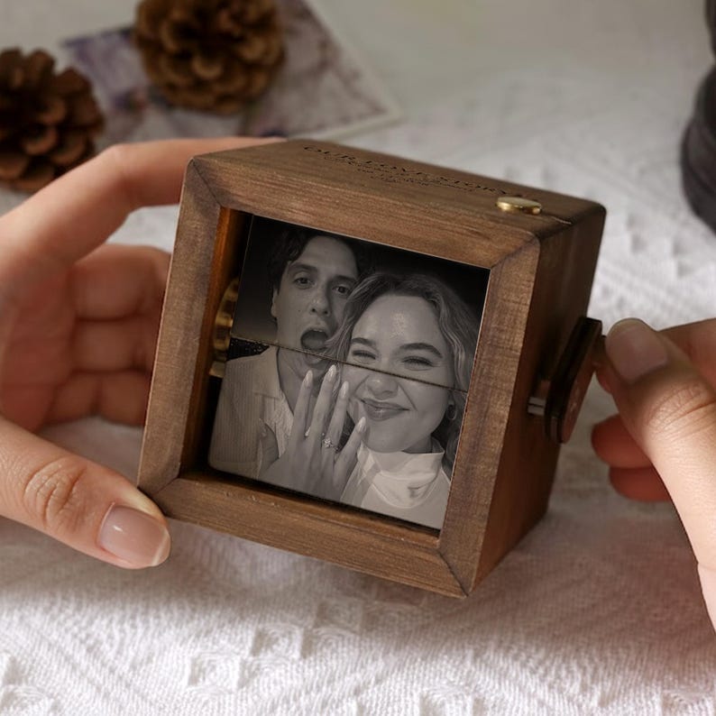 May include: A wooden photo cube with a black and white image of a couple. The cube has a crank on the side and a small gold-colored accent on top. The image is split into sections, creating a unique visual effect.