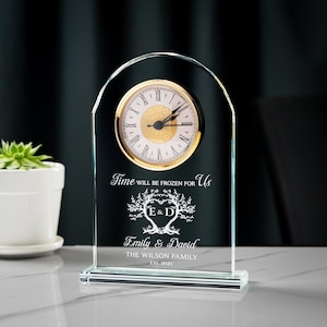 May include: A clear glass clock with a gold-colored clock face and Roman numerals. The clock has a curved top and a rectangular base. Text on the clock reads "Time will be frozen for us," with names and a date below. A decorative item.