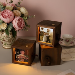 May include: Three wooden photo cubes with illuminated pictures, stacked on a table. The cubes have a crank and the text "OUR LOVE STORY". A vase of pink and white flowers and a teacup are in the background.