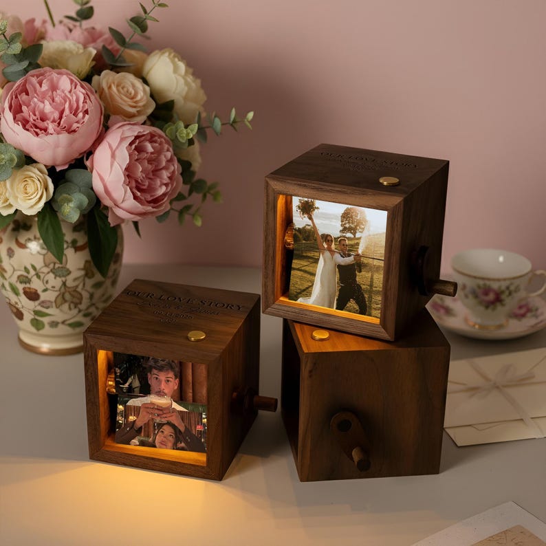 May include: Three wooden photo cubes with illuminated pictures, stacked on a table. The cubes have a crank and the text "OUR LOVE STORY". A vase of pink and white flowers and a teacup are in the background.
