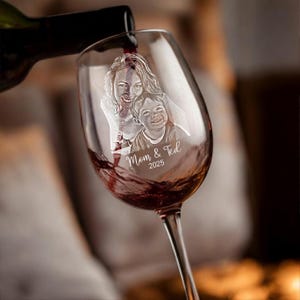 Pode incluir: Uma ta&ccedil;a de vinho transparente com um design gravado personalizado de uma m&atilde;e e um filho, com o texto "Mom & Ted 2025". O vinho tinto est&aacute; sendo derramado na ta&ccedil;a de uma garrafa escura. A ta&ccedil;a tem uma haste longa.
